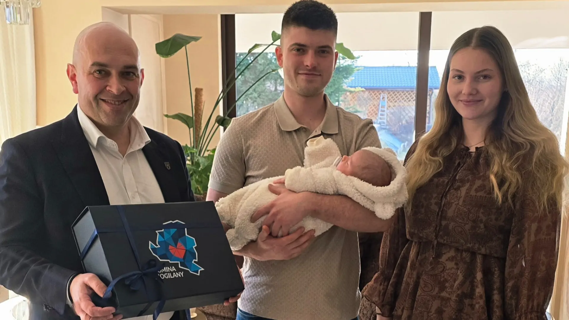 3 osoby pozują ustawione przewodem do obiektywu. Mężczyzna w środku trzyma na rękach małe dziecko, mężczyzna po lewej stronie trzyma w ręku prezentowe pudełko. Wszyscy się uśmiechają.